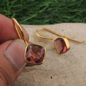 May include: A pair of gold-colored drop earrings. Each earring features a square, faceted, reddish-brown gemstone set in a gold-colored frame. The earrings are displayed against a neutral background, showcasing their design.