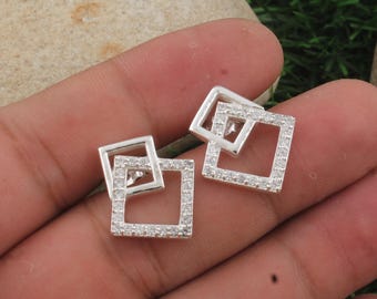 May include: A pair of silver-toned square stud earrings. Each earring features two overlapping squares, one slightly smaller than the other, encrusted with small, clear gemstones. The earrings are displayed against a neutral background.