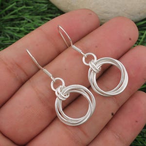 May include: A pair of silver earrings featuring a circular design. Each earring has a loop at the top with a wire hook and a cluster of interconnected silver rings. The earrings are displayed against a green background.