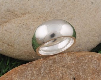 May include: A polished, silver-colored ring with a rounded top and a smooth, reflective surface. The ring is set against a neutral, textured background, highlighting its simple, elegant design. The ring's band is visible, adding to its classic appeal.