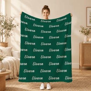 May include: A dark green blanket featuring a repeating pattern of the name Elowen printed in a white cursive script. The text is arranged in multiple horizontal rows across the entire surface of the fabric, creating a consistent design.