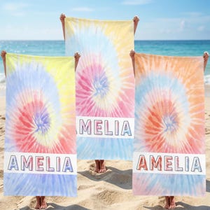 May include: Three tie-dye beach towels in shades of yellow, pink, orange, and blue are held up on a beach. Each towel has a white rectangular band with a name printed in a bold font. Two towels say "AMELIA" and one says "MELIA".