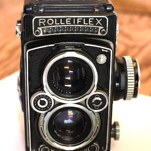 May include: A black Rolleiflex twin-lens reflex camera featuring two prominent circular lenses stacked vertically. The front panel displays the brand name ROLLEIFLEX in white text, along with various silver-toned adjustment dials, knobs, and the text Synchro-Compur and Xenotar 1:3.5/75.