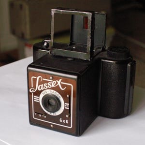 May include: A black and brown vintage film camera with a viewfinder and a lens. The camera has the text "SasseX SAS - SIAGON 6x6" on the front.
