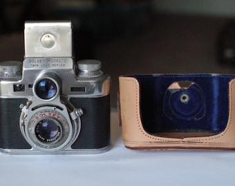 May include: A silver and black vintage twin lens reflex camera with a leather case. The camera has a lens cap and the text "BOLSEY MODEL C TWIN LENS REFLEX" on the front.