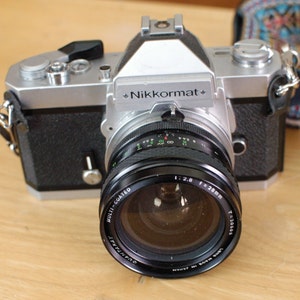 May include: A silver and black 35mm film camera with a Nikkormat lens. The lens is a 50mm f/2.8 and has a multi-coated lens.