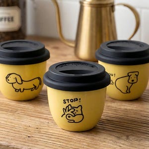 May include: Three yellow ceramic coffee cups with black lids and dog illustrations. One cup has the word "STOP!" and a dog graphic. A gold-colored kettle and a jar of coffee beans are in the background.