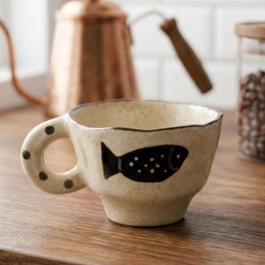 Puede incluir: Una taza de cerámica blanca con asa negra y el texto "But First, Coffee" en negrita negra. La taza está sobre un fondo borroso. La taza mide aproximadamente 10 cm de altura.