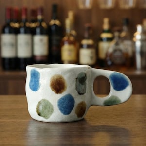Puede incluir: Una taza de cerámica hecha a mano con base blanca y lunares de colores azul, marrón y verde. La taza tiene una forma orgánica única y un asa redondeada. El fondo presenta un bar con botellas y vasos.