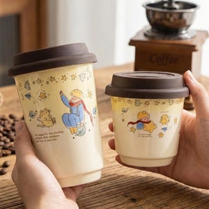 May include: Two cream-colored ceramic travel mugs with brown lids. Each mug features a cartoon illustration of a character with a red scarf and a cat, along with stars and text. A coffee grinder is in the background.