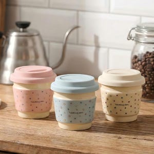 May include: Three ceramic travel mugs with pastel-colored lids and star patterns. The mugs have a cream-colored base and a matching band with the text "Twinkling stars". The lids are pink, blue, and cream.
