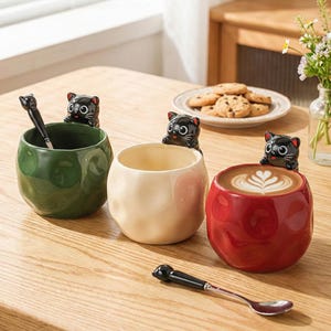 Può includere: Tre tazze in ceramica verde, crema e rossa, ciascuna con una statuina di gatto nero sul bordo. Una tazza contiene un cucchiaio e un'altra ha un latte con un disegno a cuore. Un cucchiaio è appoggiato sul tavolo.