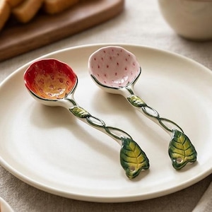 May include: Two decorative spoons with strawberry-shaped bowls, one red and orange, the other pink. The handles are silver-colored with green leaf accents. The spoons rest on a white plate, with toast and coffee in the background.
