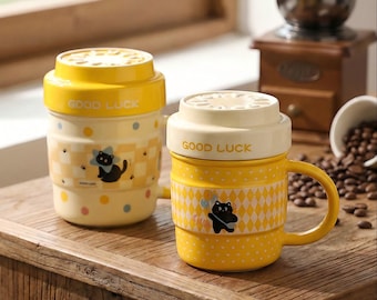 Op de afbeelding: Twee gele keramische mokken met deksels, met een zwarte kattenillustratie en de woorden "GOOD LUCK". Eén mok heeft een handvat. De mokken hebben een geruit en gestippeld patroon. Een koffiemolen en koffiebonen staan op de achtergrond.