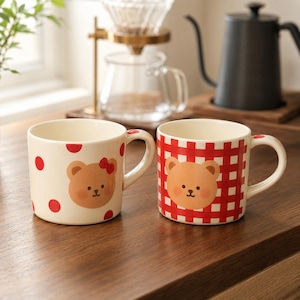 Puede incluir: Dos tazas de cerámica con motivo de oso. La taza de la izquierda es de color crema con lunares rojos y una cara de oso con un lazo rojo. La taza de la derecha presenta un patrón de cuadros rojos y blancos con una cara de oso.