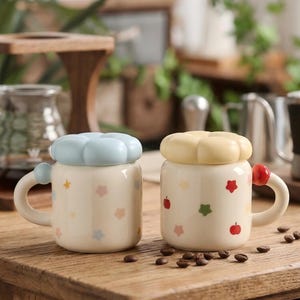 May include: Two ceramic mugs with lids, decorated with colorful star and apple designs. One mug has a light blue lid, the other a yellow lid. Both mugs have a handle and are set on a wooden surface.