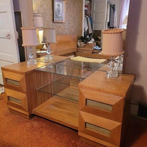 Puede incluir: Un tocador de madera vintage con un espejo grande, estantes de vidrio y dos lámparas con pantallas rosas con lunares. El tocador tiene dos juegos de cajones con diseños geométricos. El espejo refleja un armario con ropa.