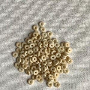May include: A pile of small, off-white, ring-shaped beads. The beads are uniform in size and shape, with a hole in the center of each. They are scattered on a textured, off-white surface.