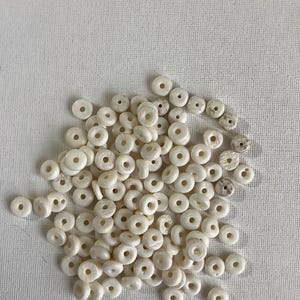 May include: A collection of small, off-white, round beads with a hole in the center. The beads are scattered on a white surface. Some beads have a slightly textured appearance, while others are smooth.