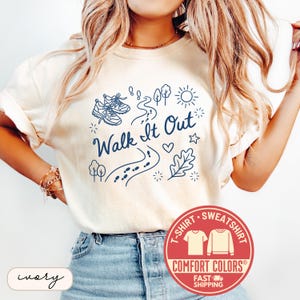 May include: Ivory-colored t-shirt with the phrase "Walk It Out" in navy blue, accompanied by illustrations of running shoes, a path, trees, and a sun. The shirt features a Comfort Colors label.
