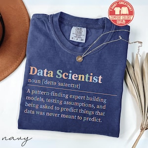 May include: Navy blue t-shirt with the words "Data Scientist" in colorful letters. The definition of "Data Scientist" is printed below the title. A gold necklace with a pendant is draped across the shirt. The shirt has a Comfort Colors tag.