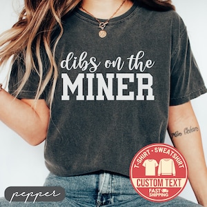 May include: Dark gray t-shirt with the text "dibs on the MINER" in white block letters. The shirt also features a red circular graphic with the words "T-SHIRT SWEATSHIRT CUSTOM TEXT FAST SHIPPING".