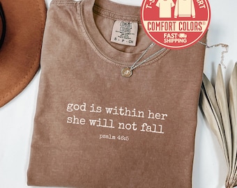 May include: Espresso-colored t-shirt with the text "god is within her she will not fall psalm 46:5" in white. The shirt has a crew neck and short sleeves. A silver necklace with a pendant is on the shirt. A red and white circular logo is in the top right corner.