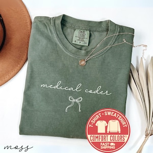 Puede incluir: Una camiseta Comfort Colors verde musgo con las palabras "medical coder" en escritura blanca y un gráfico de lazo. También se ven un collar dorado y un sombrero marrón. Una pegatina roja con "Comfort Colors" y "Fast Shipping" está en la imagen.