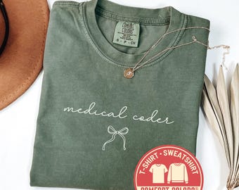 May include: A moss green Comfort Colors t-shirt with the words "medical coder" in white script and a bow graphic. A gold necklace and a brown hat are also visible. A red sticker with "Comfort Colors" and "Fast Shipping" is in the image.