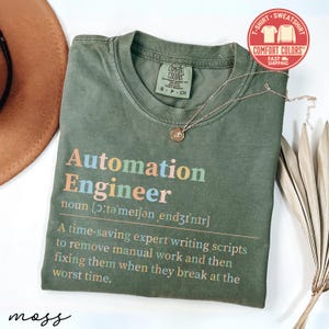May include: Olive green t-shirt with the text "Automation Engineer" in colorful letters. The shirt includes a definition and description of the job. A gold necklace with a pendant rests on the shirt.