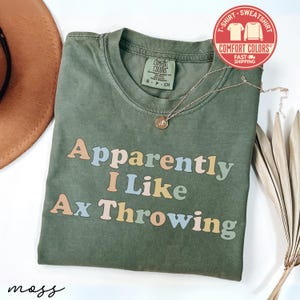 May include: An olive green t-shirt with the phrase "Apparently I Like Ax Throwing" in pastel colors. A gold necklace with a pendant rests on the shirt. The shirt has a Comfort Colors tag.