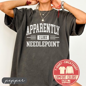 May include: Dark grey t-shirt with the text "APPARENTLY I LIKE NEEDLEPOINT" in white block letters. The shirt has a crew neck and short sleeves. A red and white circular logo is visible on the bottom right.