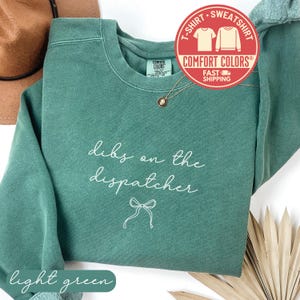 May include: Light green sweatshirt with the text "dibs on the dispatcher" in white script. A bow graphic is below the text. The sweatshirt has a crew neck and a Comfort Colors tag.
