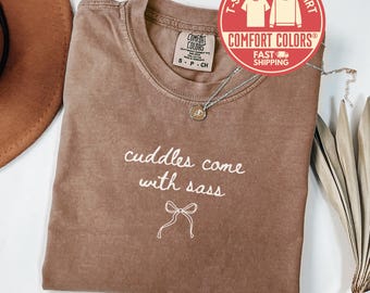 May include: A brown Comfort Colors t-shirt with the phrase "cuddles come with sass" in white script. A small bow graphic is below the text. The shirt has a crew neck and short sleeves. A silver necklace with a pendant is on the shirt.
