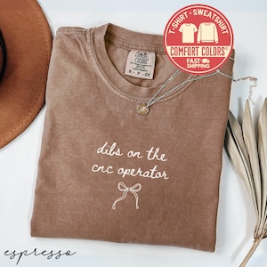 May include: Espresso-colored t-shirt with the phrase "dibs on the cnc operator" in white script, along with a bow graphic. The shirt has a Comfort Colors label and a silver necklace. A brown hat and dried plant are in the background.