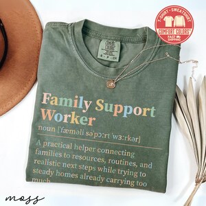 May include: Olive green t-shirt with the words "Family Support Worker" in colorful letters. The shirt includes a definition of the term. A gold necklace and a brown hat are also in the image.
