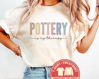 May include: Ivory-colored t-shirt with the text "POTTERY is my therapy" in pastel colors. The shirt has a crew neck and short sleeves. A red circular logo with the words "T-SHIRT SWEATSHIRT" and "COMFORT COLORS" is visible.