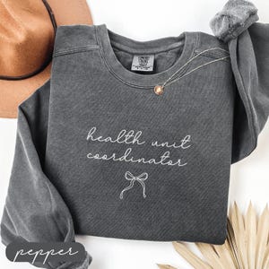 May include: A dark gray sweatshirt with the words "health unit coordinator" in white script, along with a bow graphic. A gold necklace with a pendant is draped on the shirt. A brown hat is visible in the background.