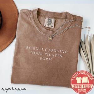 May include: Espresso-colored Comfort Colors t-shirt with the text "SILENTLY JUDGING YOUR PILATES FORM" in white. A silver necklace with a pendant rests on the shirt. The shirt has a crew neck and short sleeves. A brown hat is partially visible.