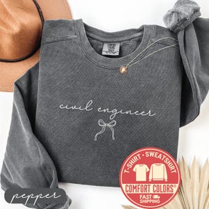 May include: A dark gray Comfort Colors sweatshirt with the words "civil engineer" in cursive and a bow graphic. A gold necklace and a brown hat are visible. The sweatshirt has a crew neck and a red and white Comfort Colors logo.