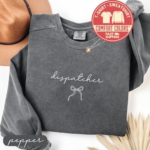 May include: A dark gray sweatshirt with the word "dispatcher" in white script and a bow graphic. The sweatshirt has a crew neck and long sleeves. A Comfort Colors tag is visible. The word "pepper" is at the bottom.