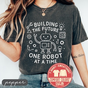 May include: Dark gray Comfort Colors t-shirt with a white graphic of a robot and the text "BUILDING THE FUTURE ONE ROBOT AT A TIME." A red and white Comfort Colors logo is visible. The shirt is a t-shirt.