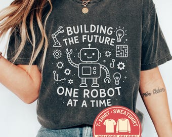 May include: Dark gray Comfort Colors t-shirt with a white graphic of a robot and the text "BUILDING THE FUTURE ONE ROBOT AT A TIME." A red and white Comfort Colors logo is visible. The shirt is a t-shirt.