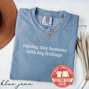 May include: A blue jean colored t-shirt with the text "raising tiny humans with big feelings" in white. A gold necklace with a pendant rests on the shirt. The shirt has a Comfort Colors tag.