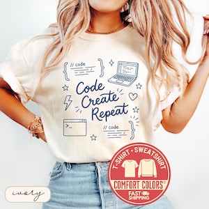 May include: Ivory-colored t-shirt with the text "Code Create Repeat" in navy blue, along with a laptop, code brackets, and stars. A red Comfort Colors logo is at the bottom. The shirt is a t-shirt and sweatshirt.