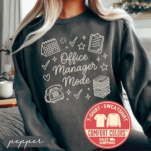 May include: A dark gray sweatshirt with the words "Office Manager Mode" in white, along with various office-related graphics. The sweatshirt has a crew neck and long sleeves. A red circular logo with the words "Comfort Colors" is visible.
