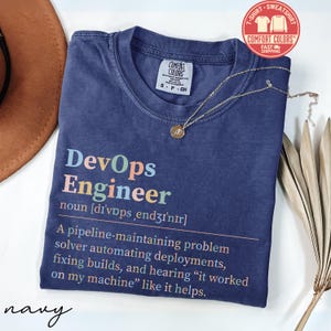 May include: Navy blue Comfort Colors t-shirt with the text "DevOps Engineer" in colorful letters. The definition of DevOps Engineer is printed below the title. A gold necklace with a pendant is draped on the shirt.