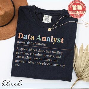May include: Black t-shirt with the text "Data Analyst" in colorful letters. The definition of a data analyst is printed below the title. A gold necklace and a brown hat are also in the image.