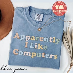 May include: A blue jean colored t-shirt with a crew neckline and a Comfort Colors brand tag. The shirt features the text "Apparently I Like Computers" printed in a multi-colored, rounded font. A thin gold chain necklace rests on the shirt.