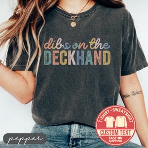 May include: Dark gray t-shirt with the text "dibs on the DECKHAND" in colorful letters. A red circle with "T-SHIRT SWEATSHIRT CUSTOM TEXT FAST SHIPPING" is in the lower right corner. The word "pepper" is in the lower left corner.